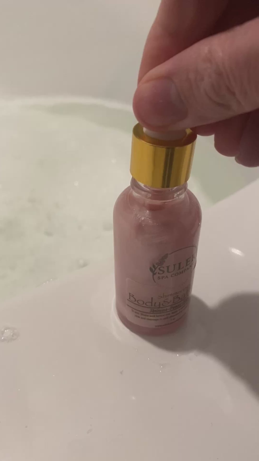 Shimmering body and bath oil bottle shown being added to bath water for a luminous, hydrated glow, Sulek Spa Company